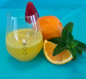 A Mimosa in a Happy Emoji Measuring Wine Glass topped with a strawberry. An orange slice and fresh mint sit next to it.