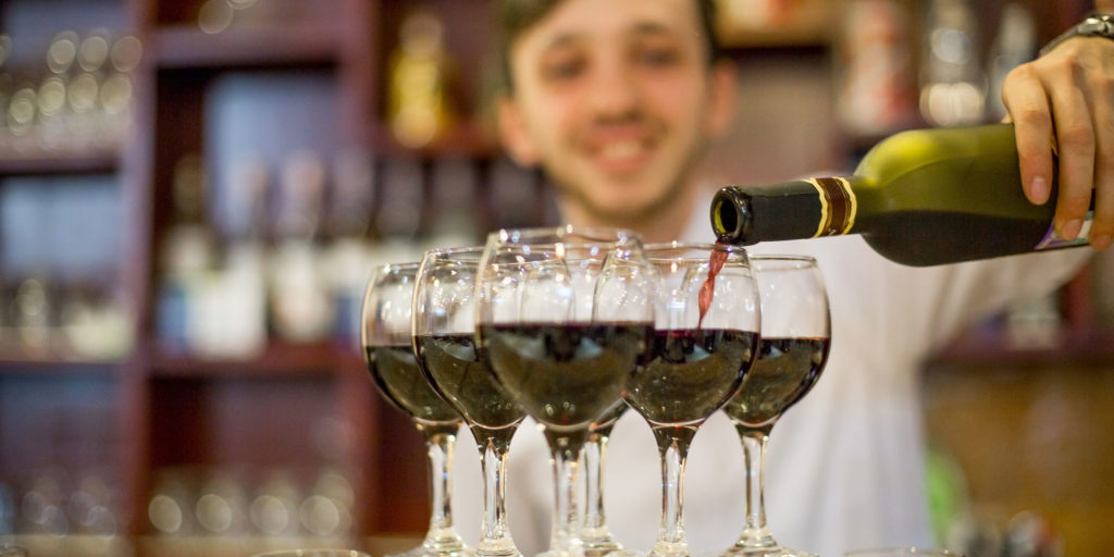 Man pouring red wine into multiple glasses at bar.